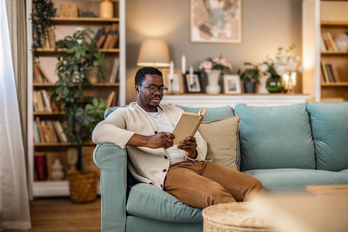 A photo of a Black man reading a book while sitting on the couch in the living room.