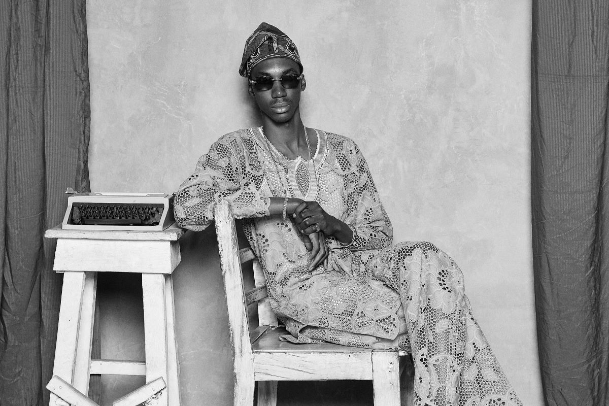 A photo of Layi Wasabi sitting on a wooden chair with a typewriter on a stool beside him.