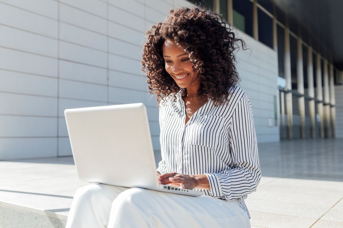 A portrait of a smiling young businesswoman using a laptop while sitting outside an office building.