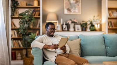 A photo of a Black man reading a book while sitting on the couch in the living room.