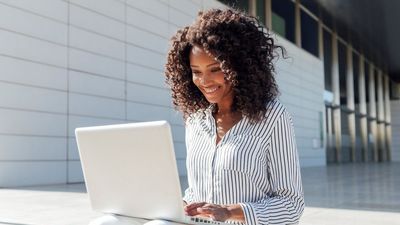 A portrait of a smiling young businesswoman using a laptop while sitting outside an office building.
