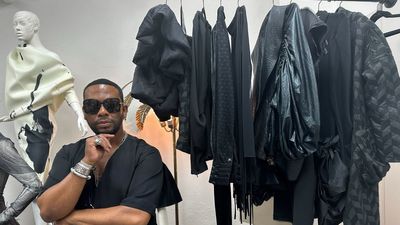 A photo of David Tlale in his New York showroom with some of his pieces on a hanger to his left.